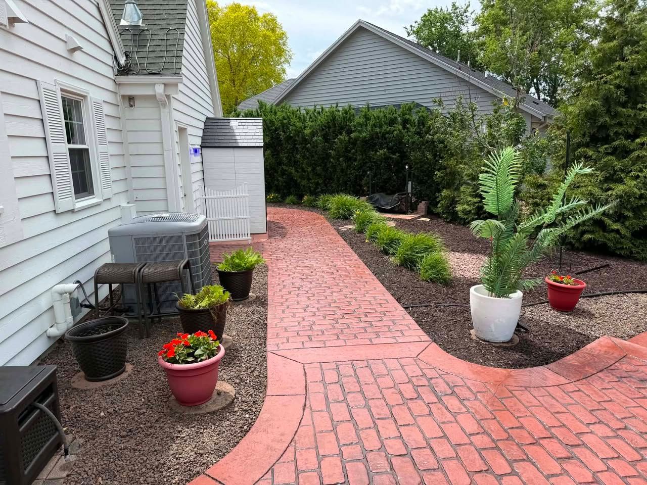 Stamped concrete garden path beside white house, with potted flowers and shrubs leading to a hedge-lined yard