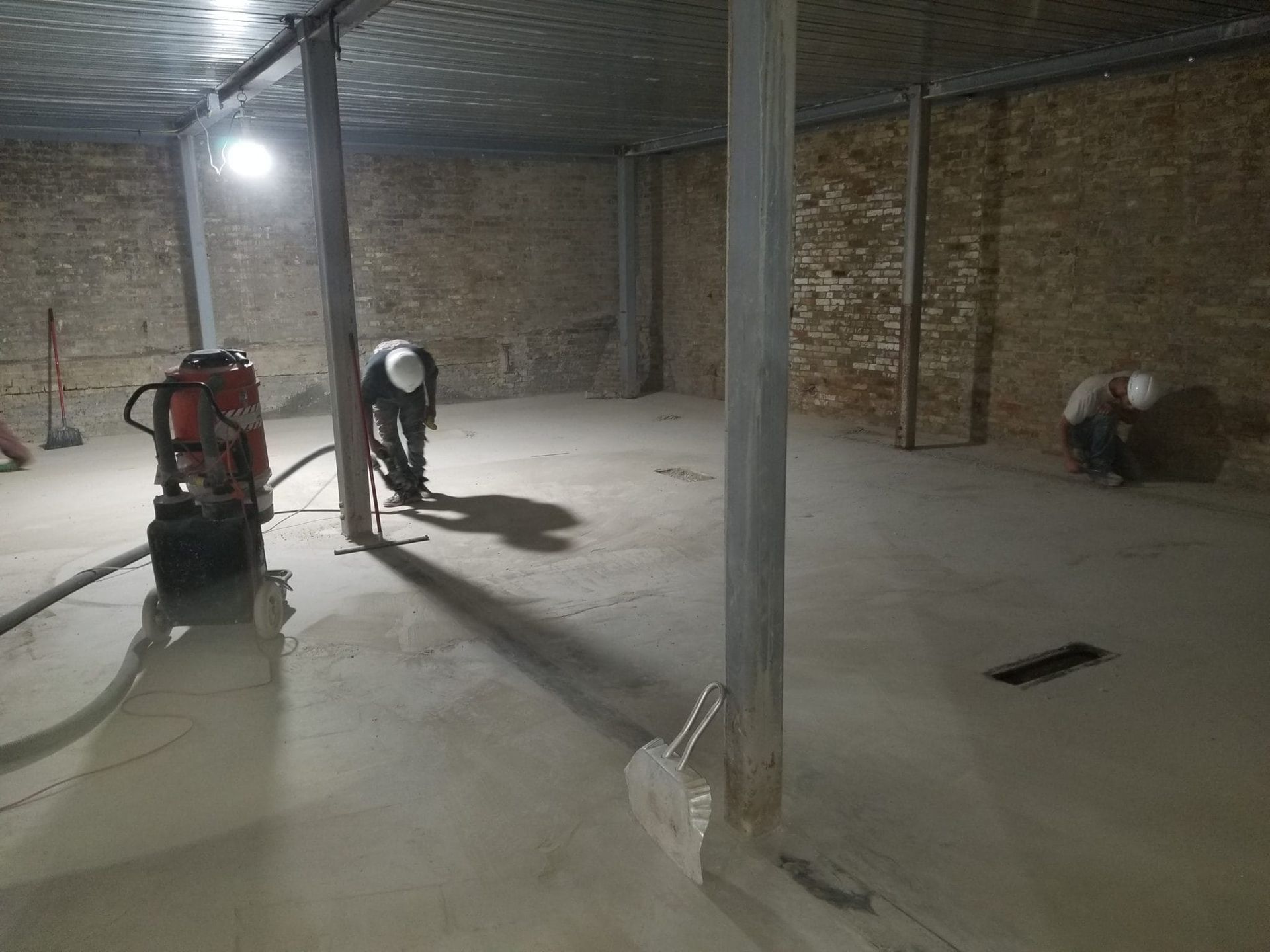 Large unfinished room with concrete floor, support columns, and workers using construction equipment before polishing concrete.
