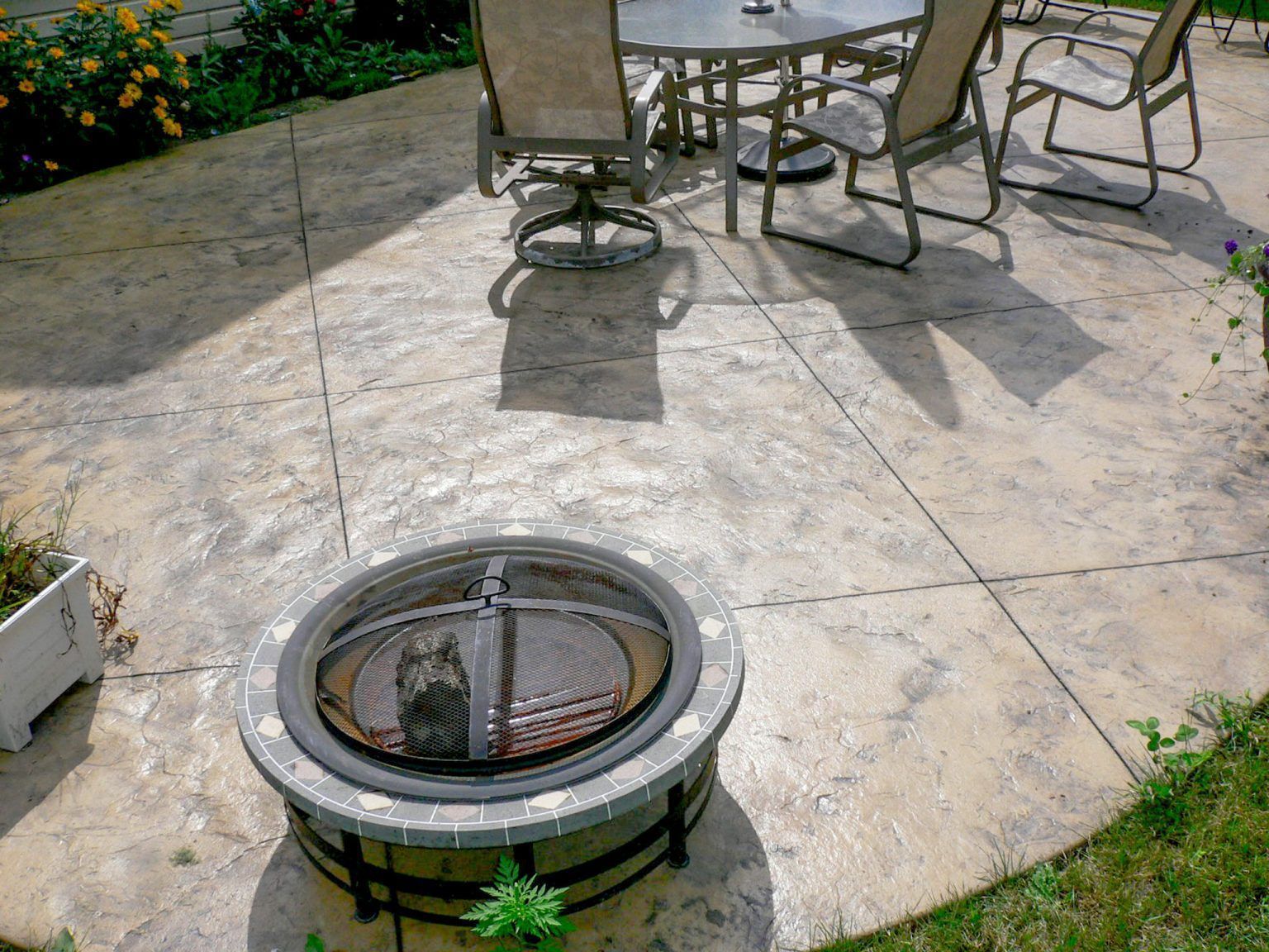 Stamped concrete patio with a round outdoor fire pit and patio chairs in bright sunlight.