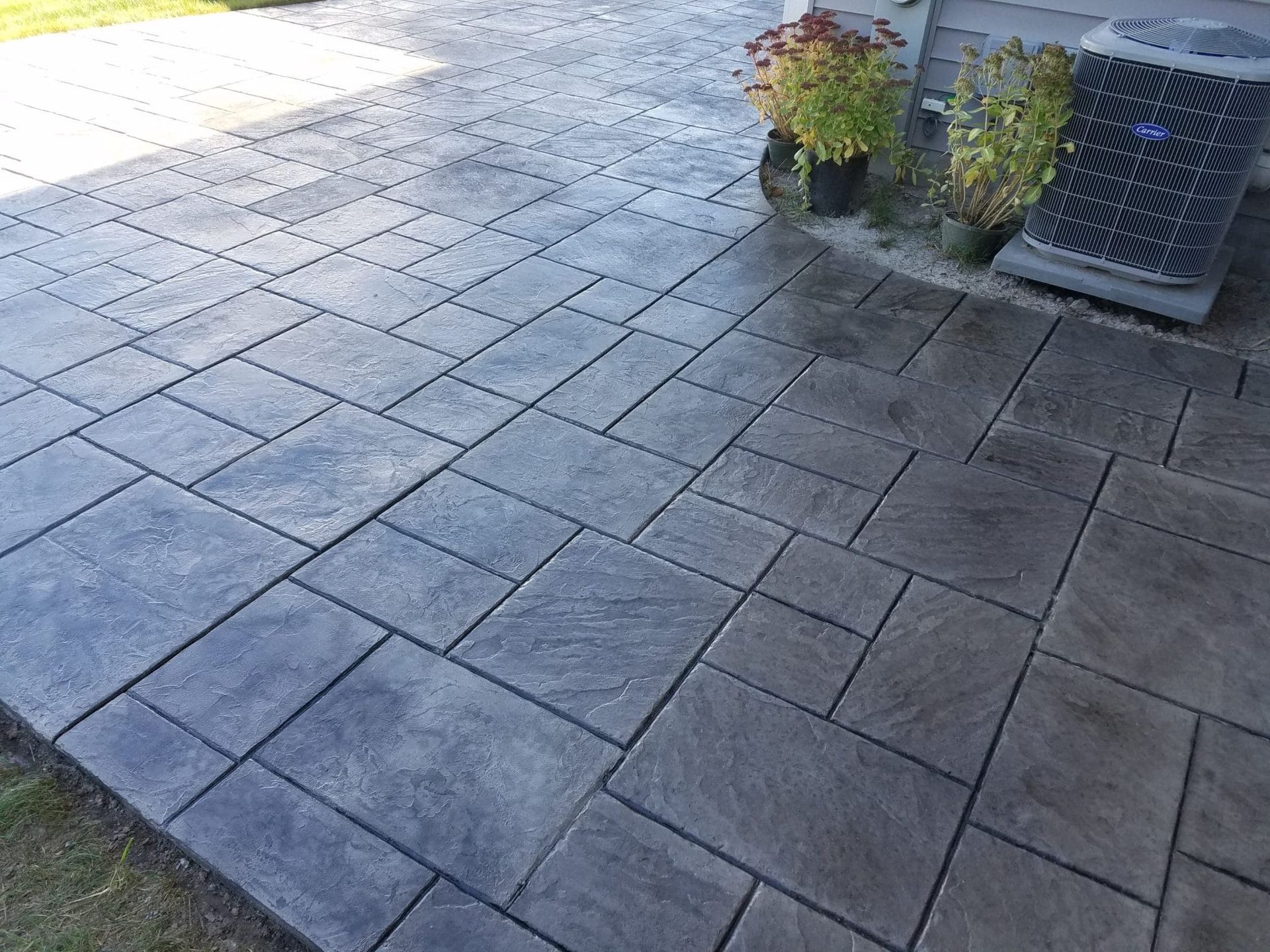 Stamped concrete patio with planters and a vent on the right, sunlight casting shadows across the tiles