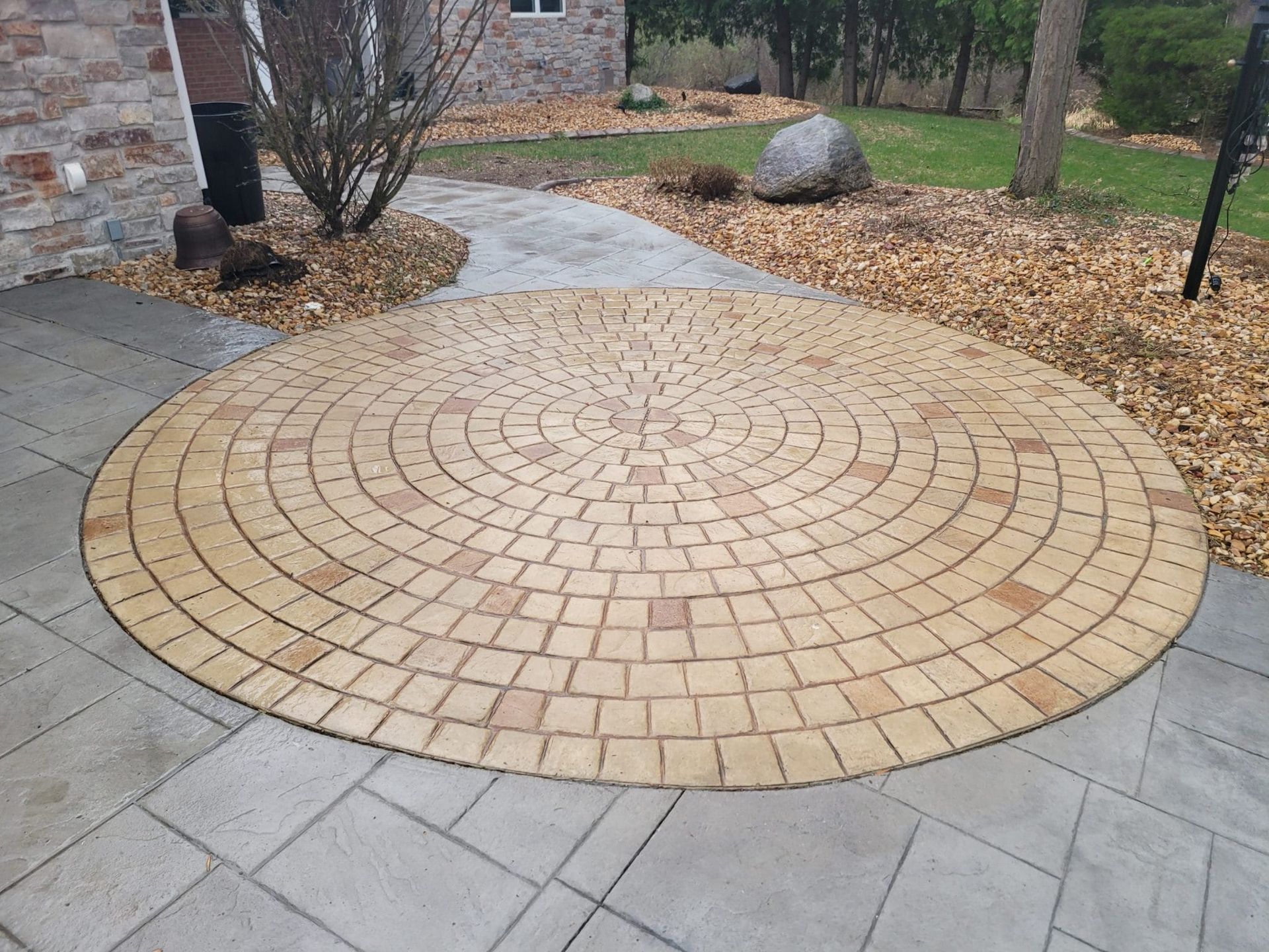 Circular stamped concrete in a landscaped yard beside a stone house and paved walkway.