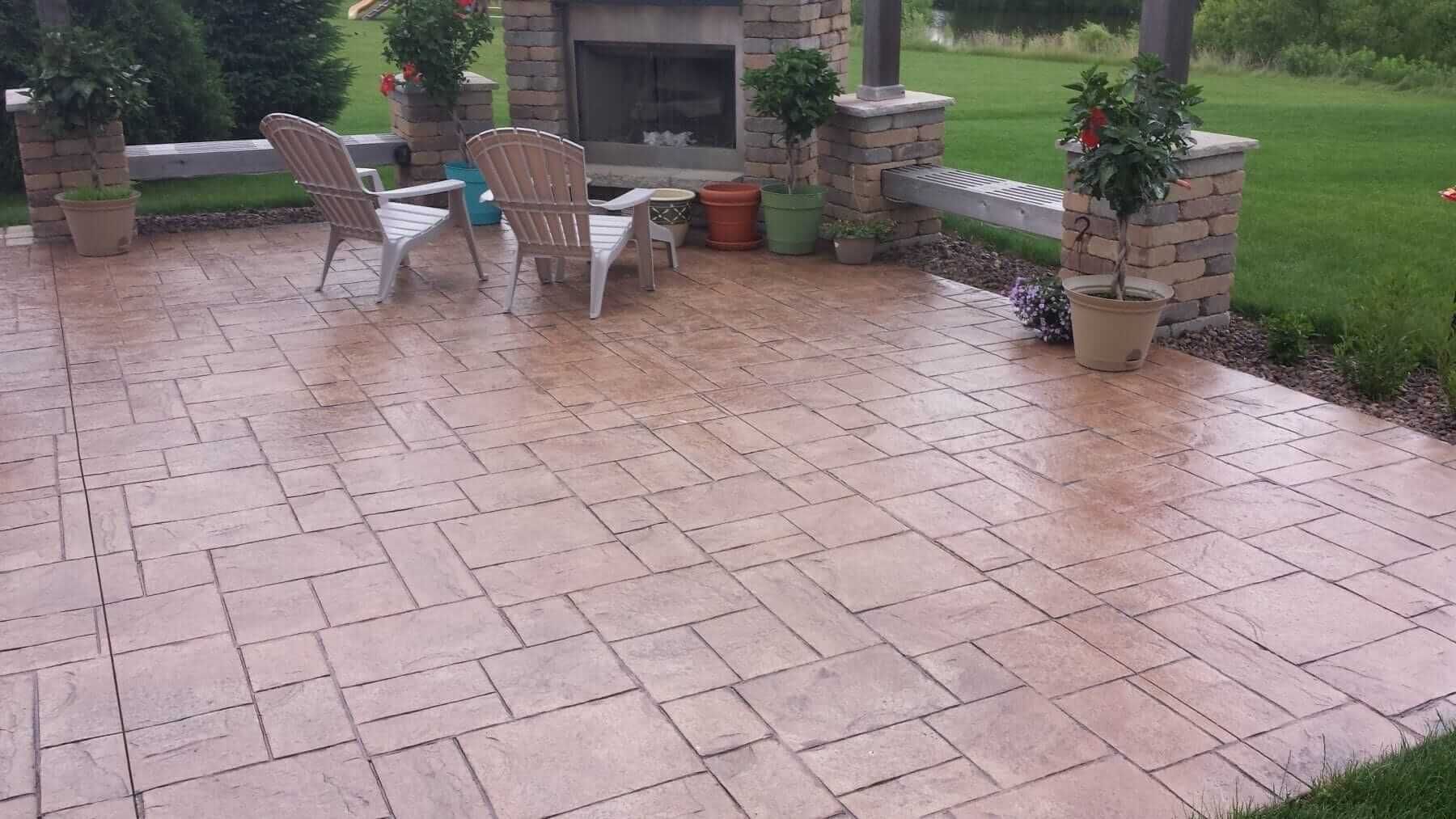 Stamped concrete patio with wet brick pavers, two chairs, and potted plants beside a garden lawn.