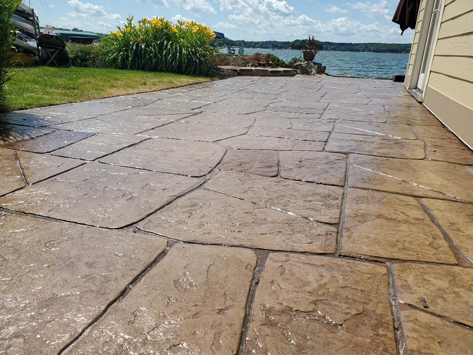 Stamped concrete patio walkway beside a house leading to a waterfront view with grassy garden and flowers