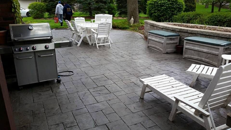 Stamped concrete patio with grill, white lounge chairs, and benches on a stone surface near a garden path