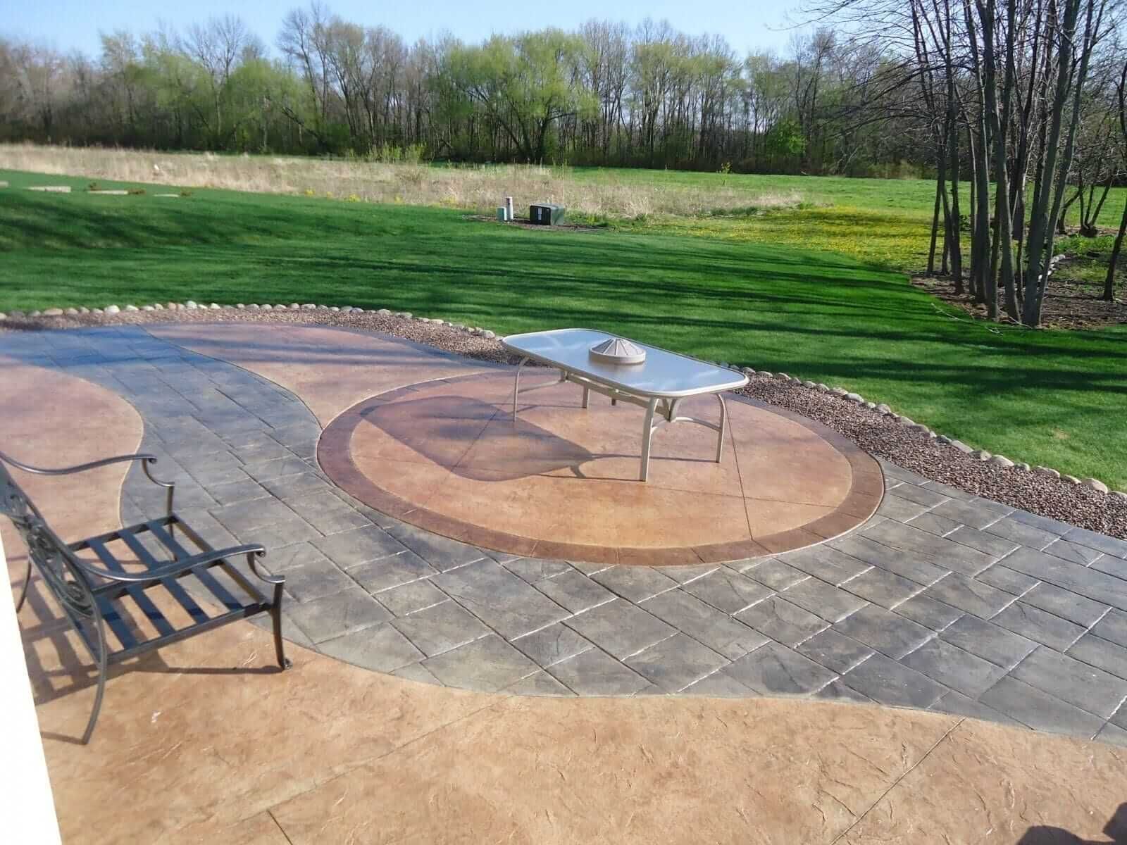 Backyard stamped concrete patio with fire pit table, chairs, and lawn beyond