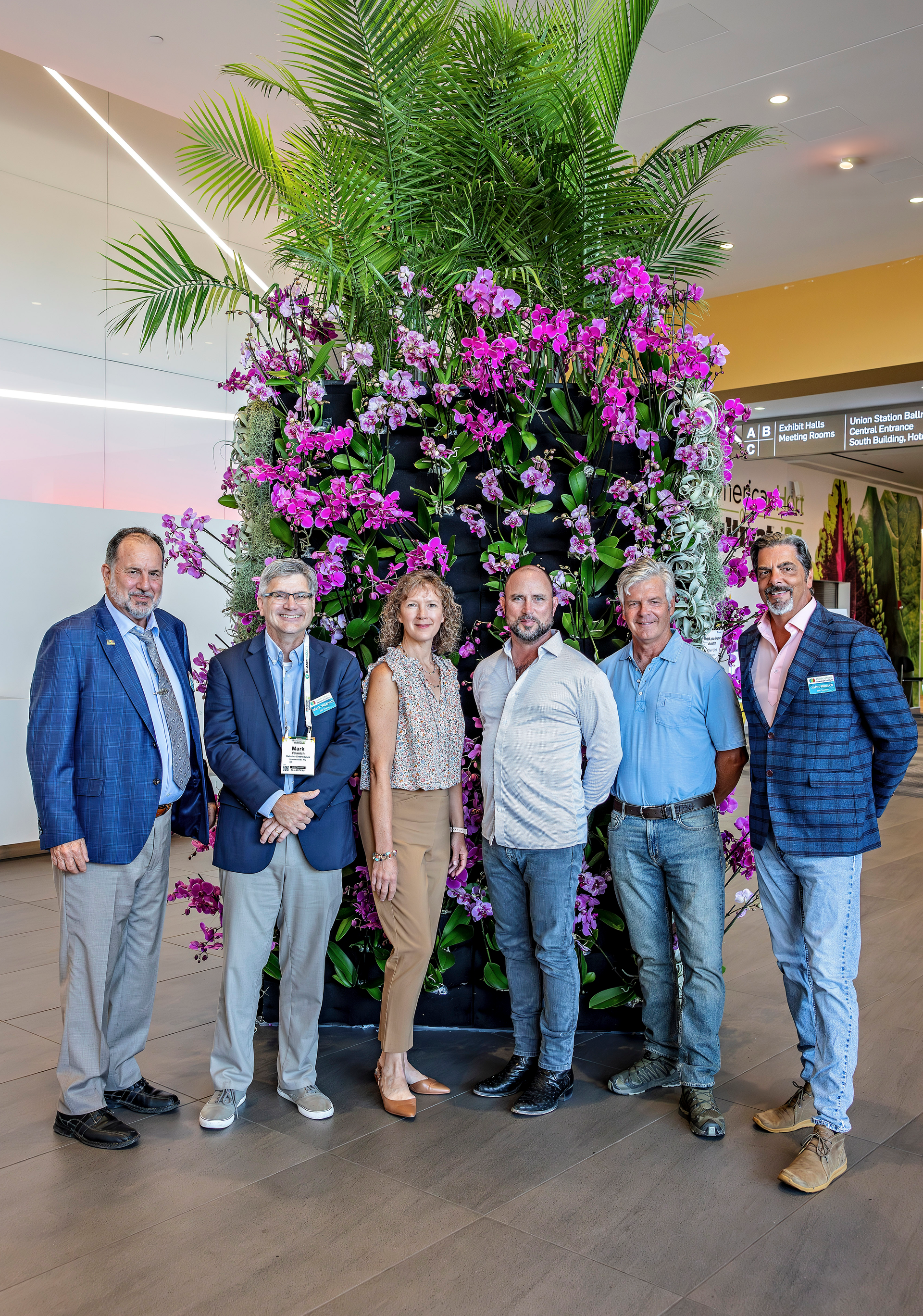 Group of six people posing in front of a large floral display, in a modern building.