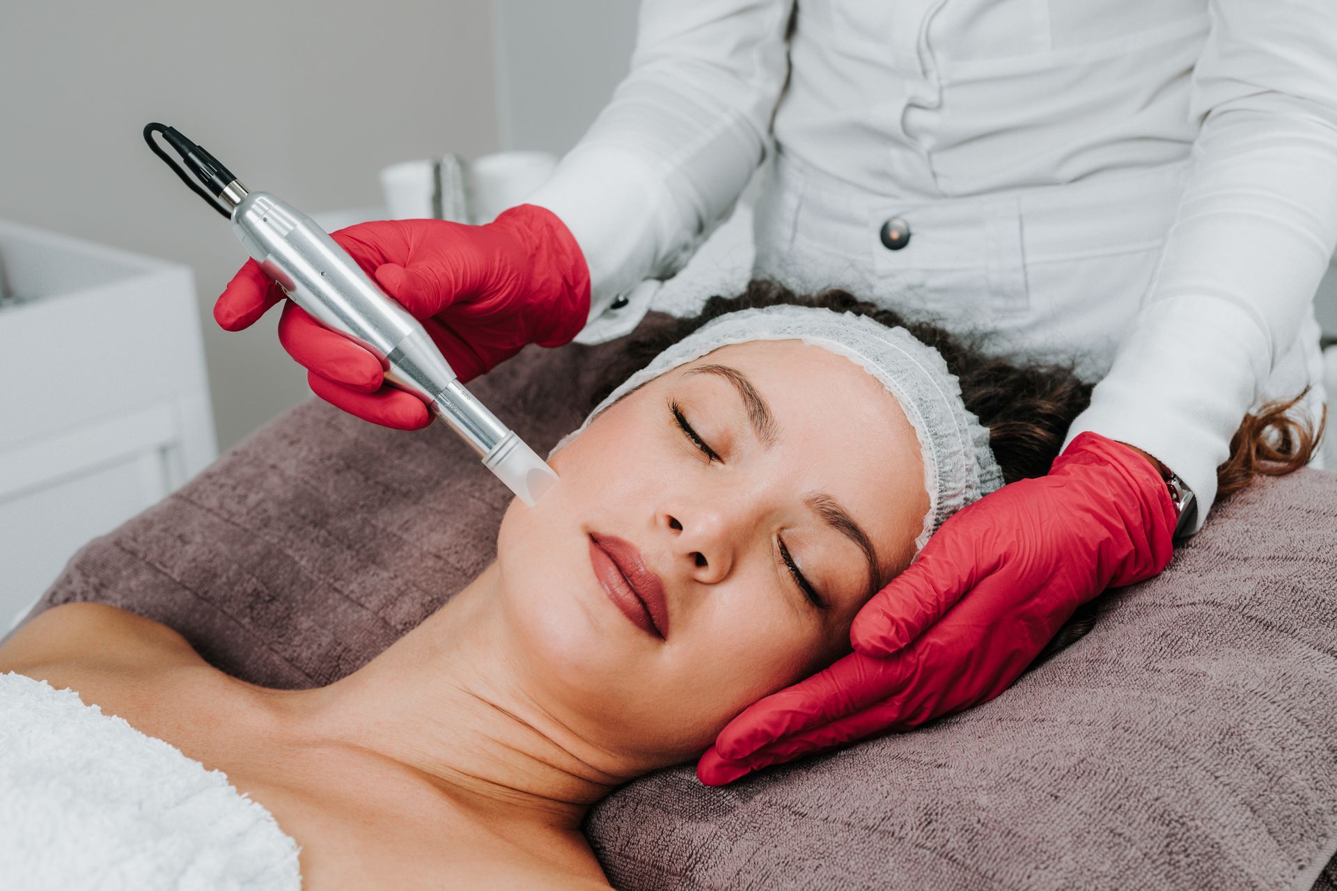 A practitioner in red gloves uses a handheld cosmetic device on a person’s face during a skin treatment.