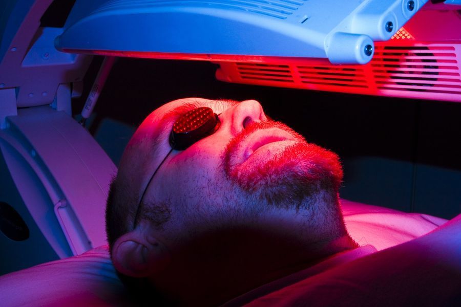 A person lies under a red light therapy panel, wearing protective eye goggles while receiving facial treatment.