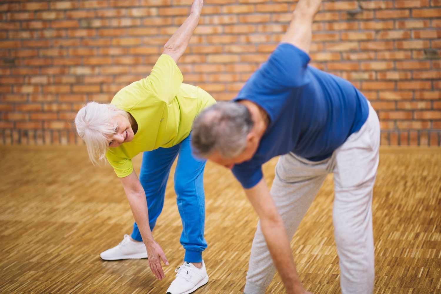 An elderly couple is doing stretching exercises together in a gym.
