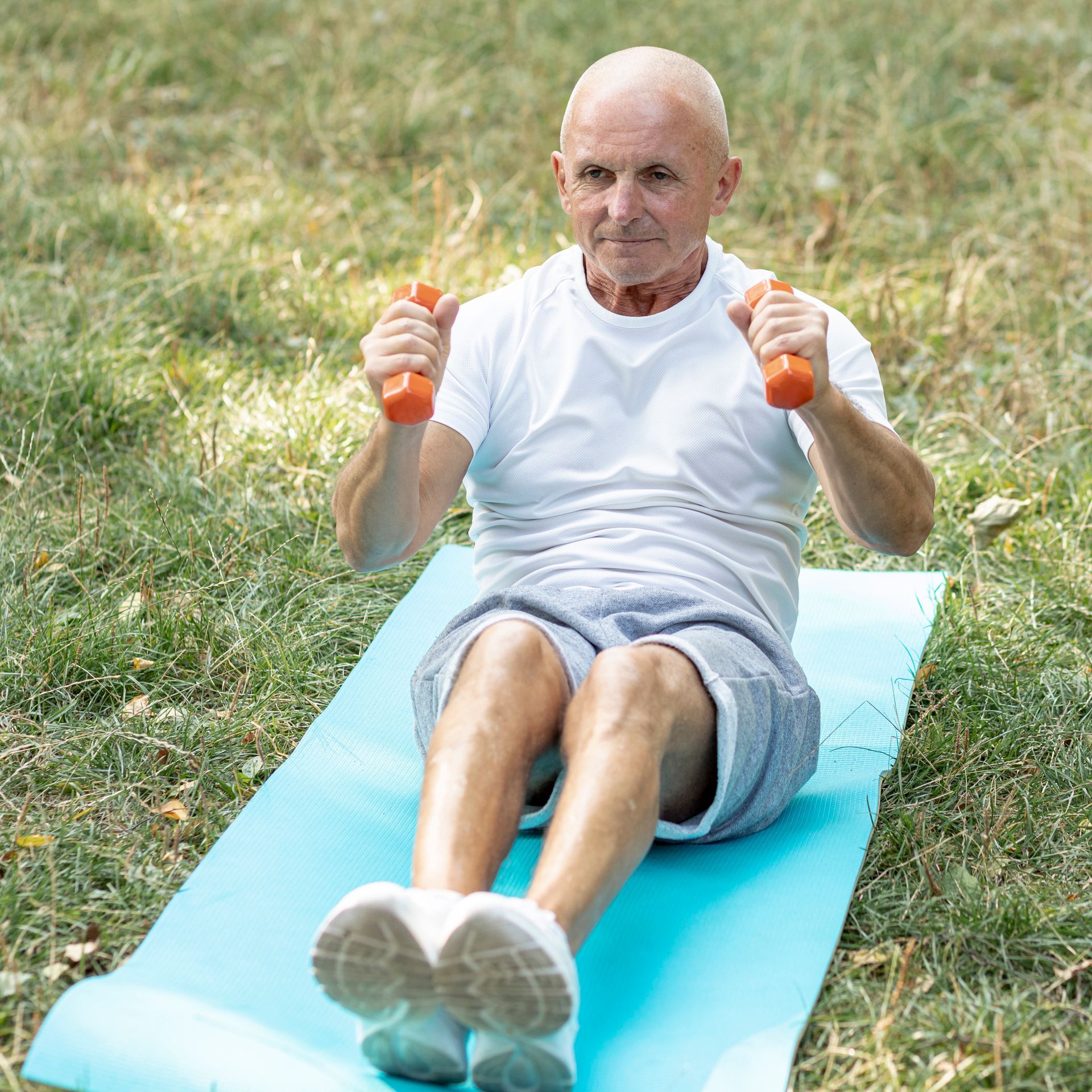 An elderly man is sitting on a yoga mat holding dumbbells.