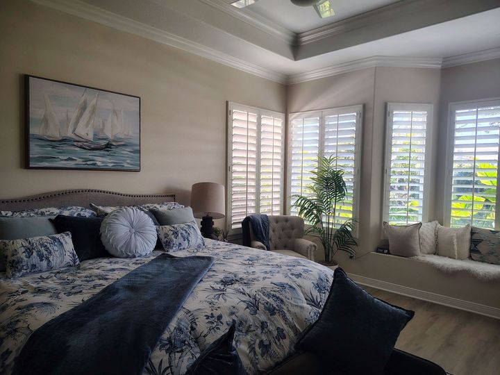 Bright bedroom with a large bed, ocean artwork, and windows with white shutters and sunlight.