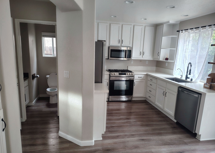 Bright kitchen with white cabinets, stainless steel appliances, and dark wood floors; hallway leads to a bathroom.