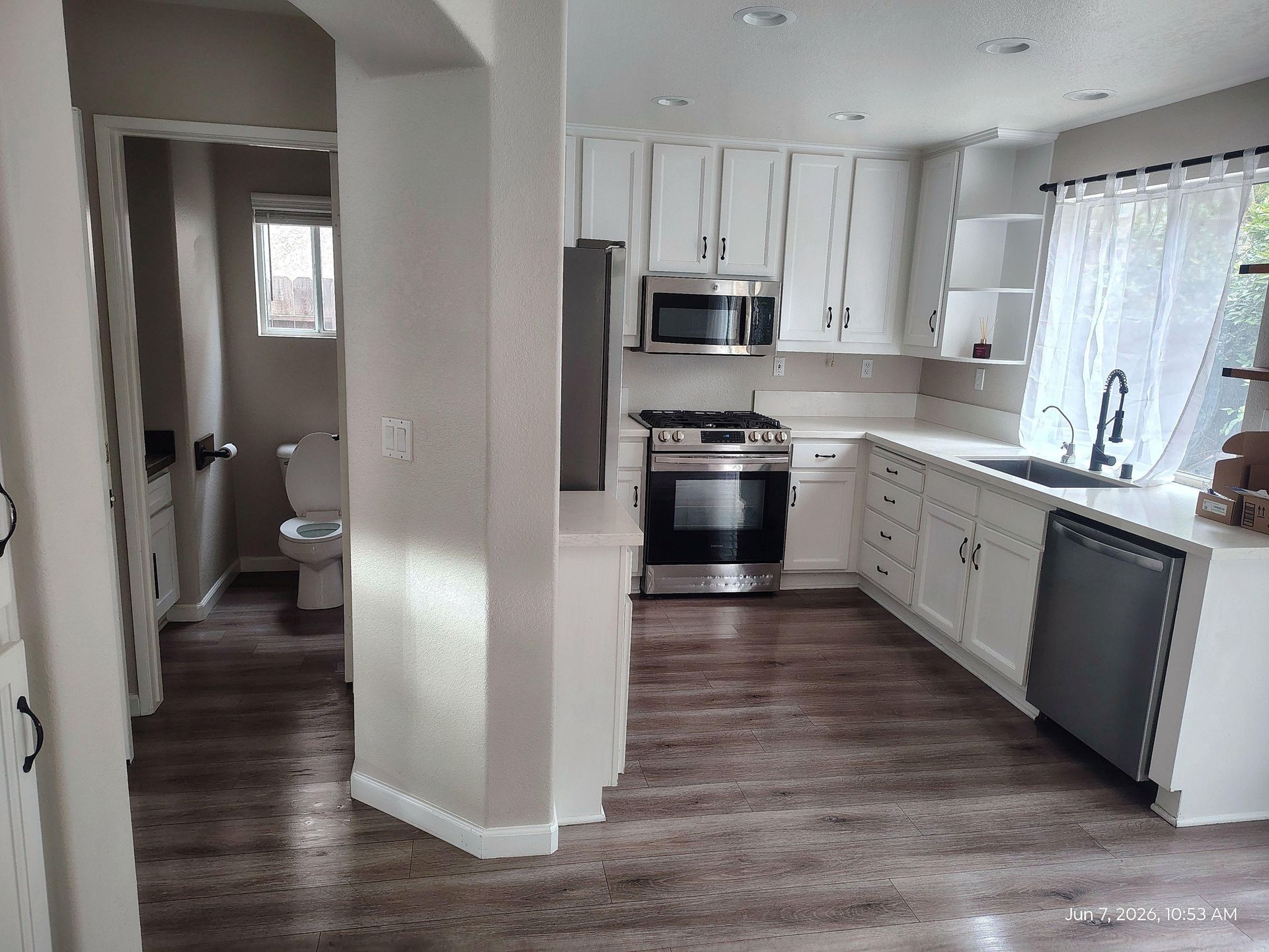 Small kitchen with white cabinets, stainless stove and microwave, dark wood floors, and a bathroom visible through doorway