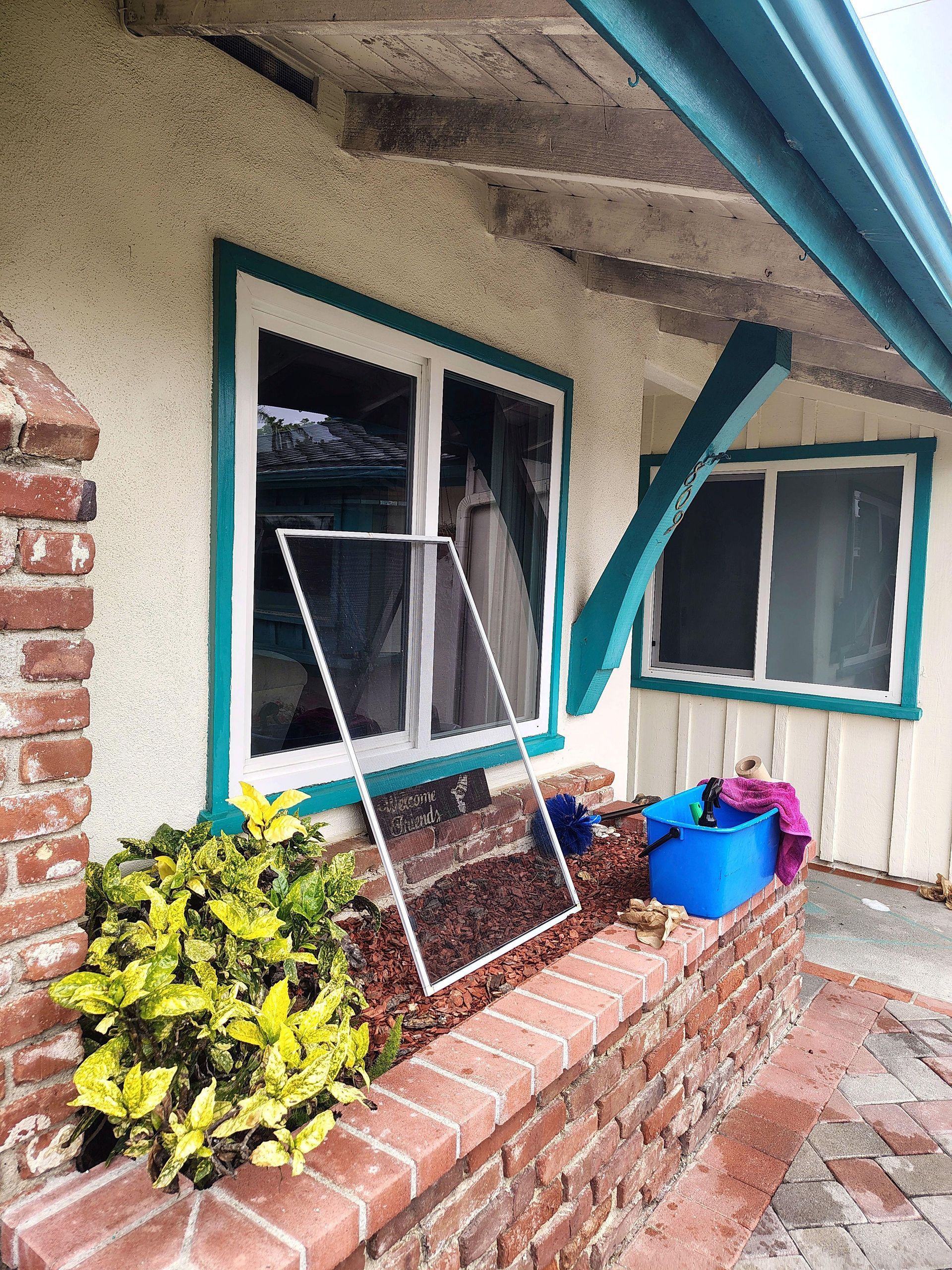 Brick porch with two windows, a leaning screen, and blue and pink buckets under a teal-trimmed roof