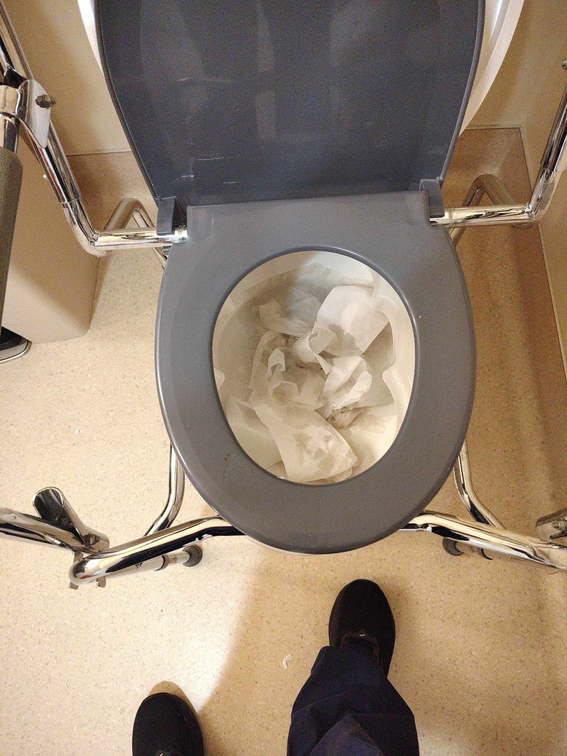 Overhead view of a toilet with crumpled tissue inside and a person’s shoe visible below.