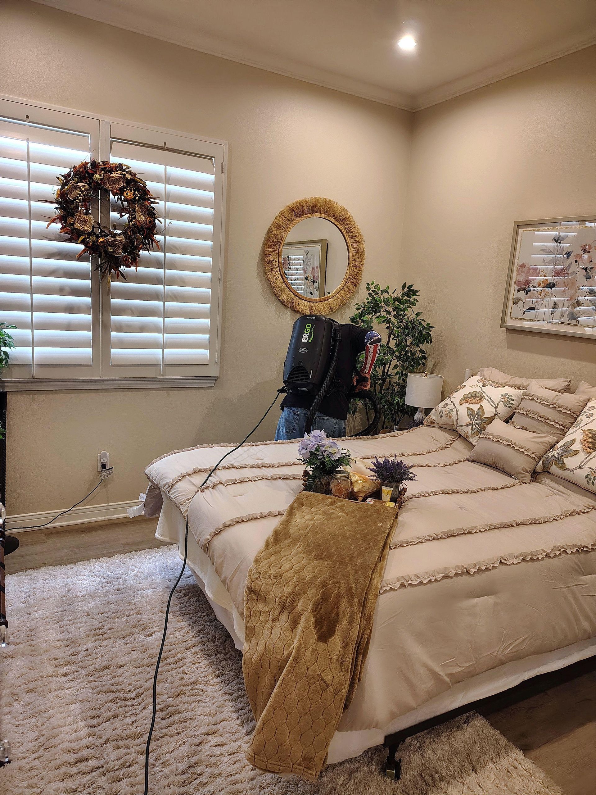 Cozy beige bedroom with a made bed, wreath on the window, mirror wall decor, and shag rug.