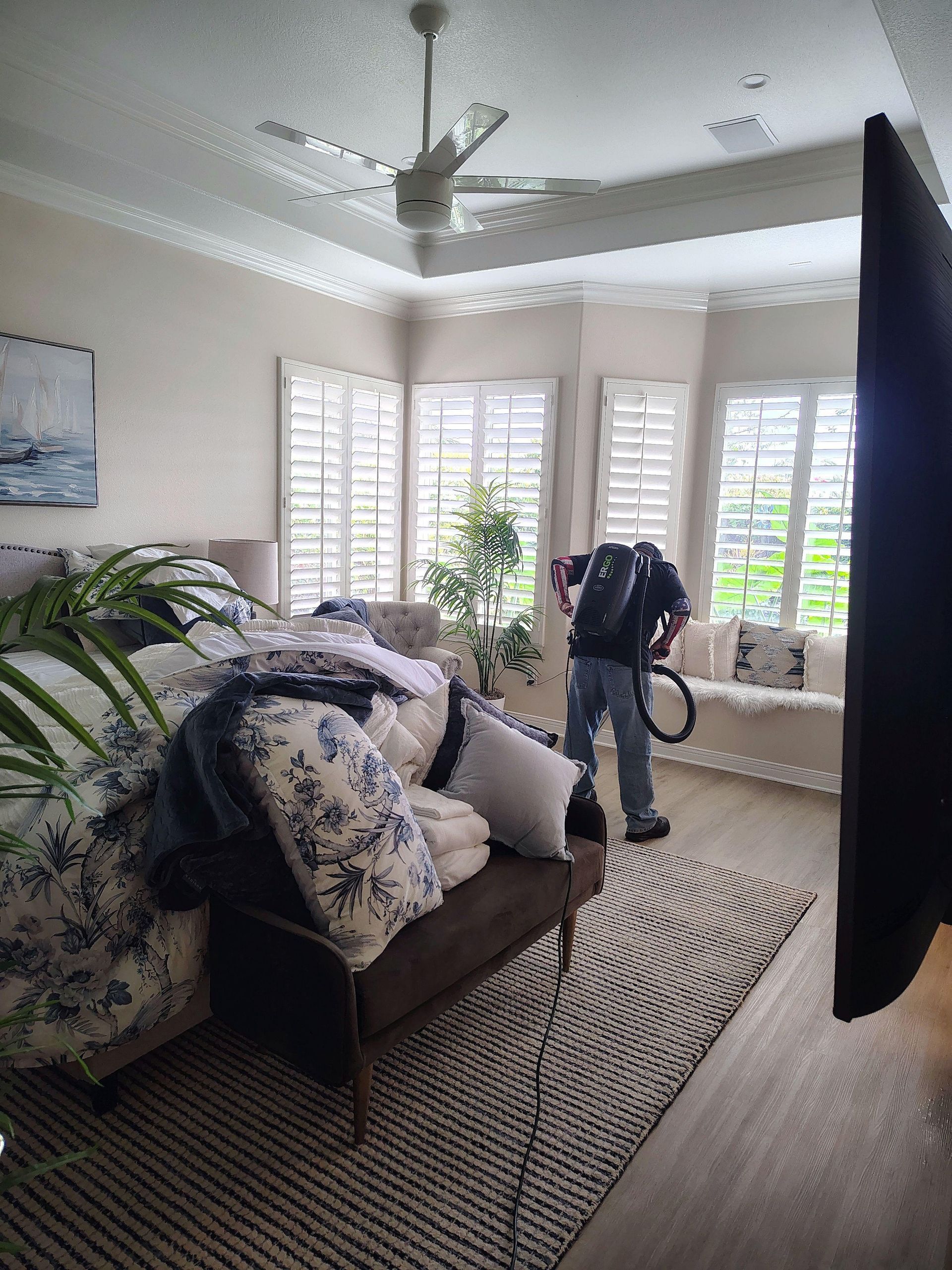 Living room with a couch, plants, shuttered windows, and a person vacuuming near the windows.