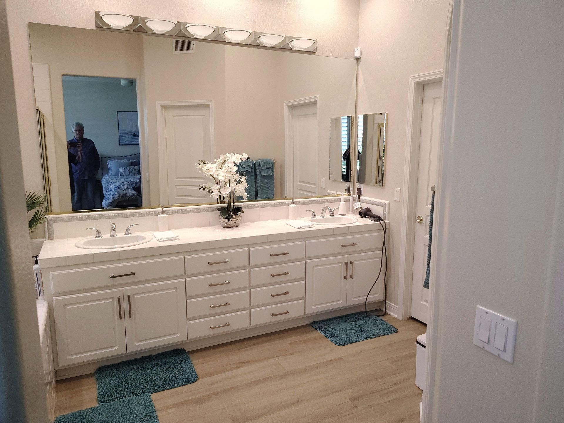 Spacious white bathroom vanity with double sinks, mirrors, and blue bath mats on a wood floor