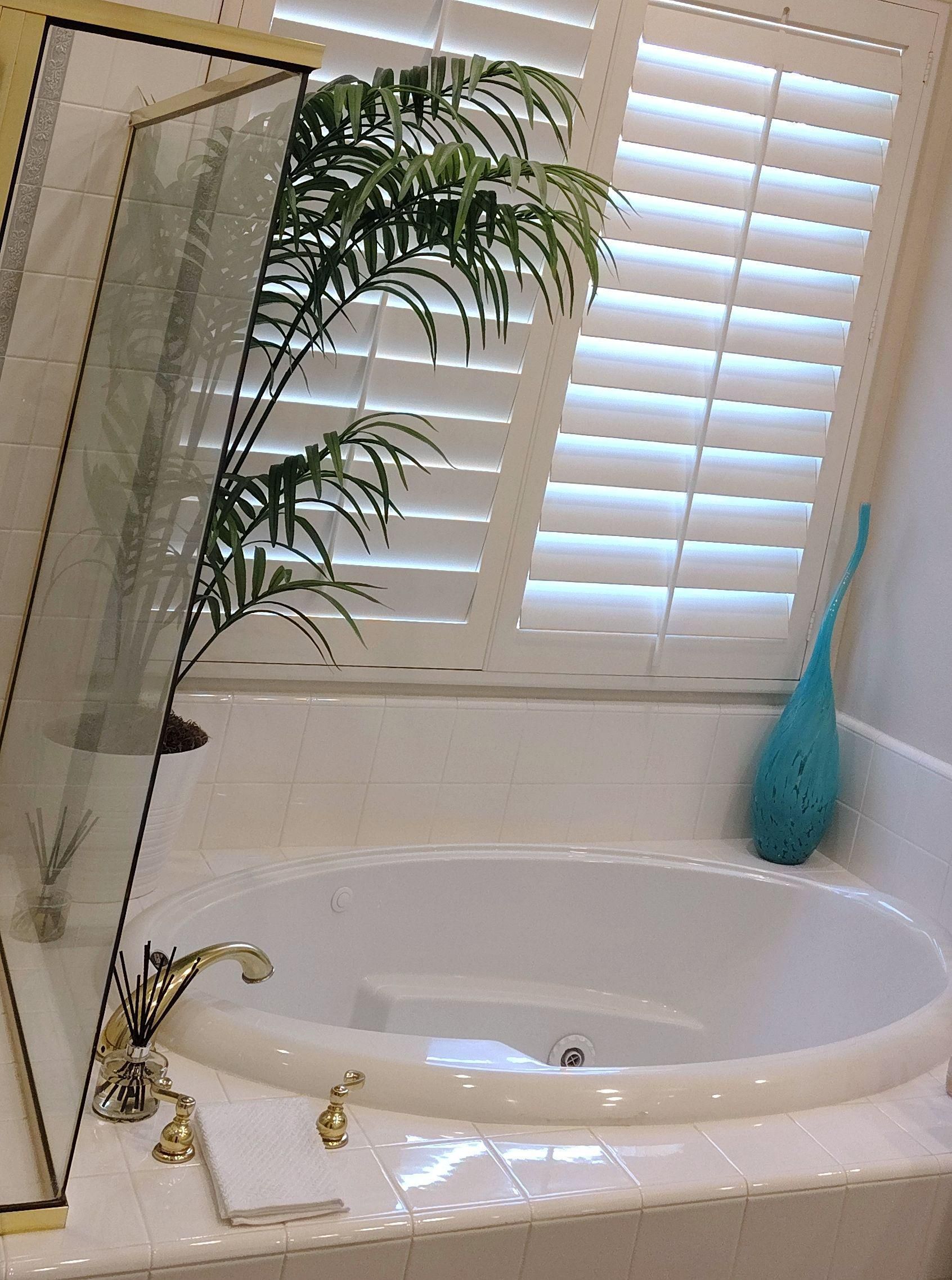 Bright bathroom with a bathtub, gold faucet, potted palm, and blue vase by shuttered window.
