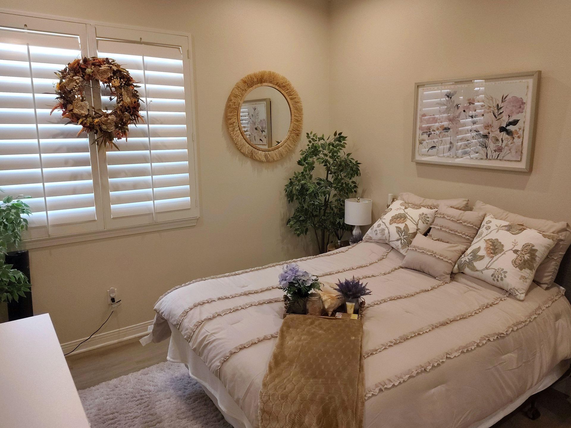 Cozy beige bedroom with a made bed, wreath on shutters, round mirror, and floral wall art.