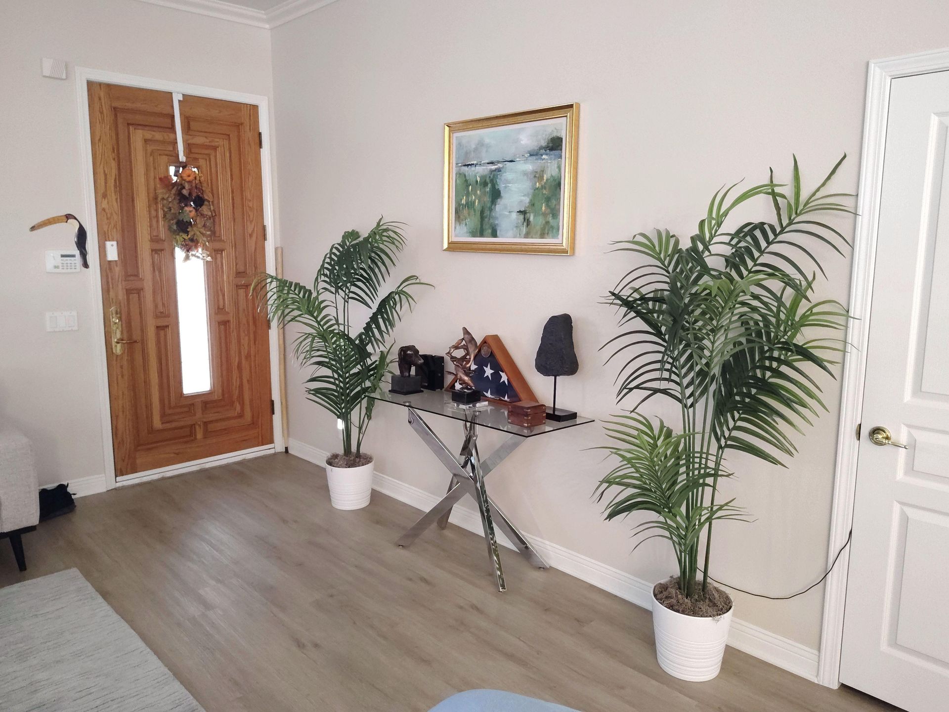Bright entryway with wooden front door, two potted palms, a small table, and framed wall art.