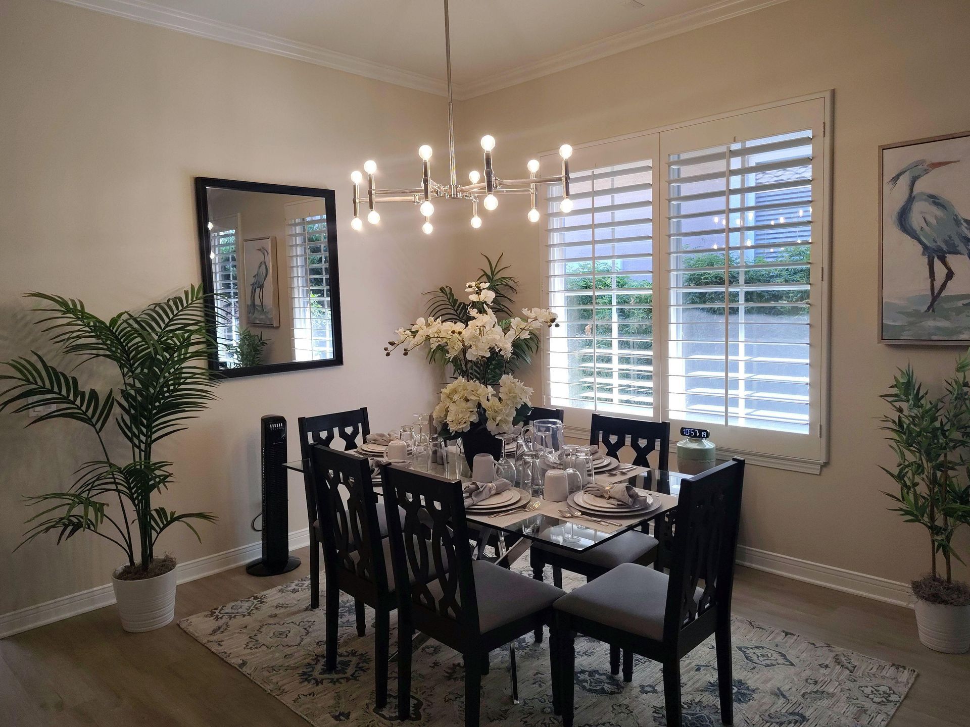 Dining room with black table and chairs, chandelier, floral centerpiece, and white shutters by a window.