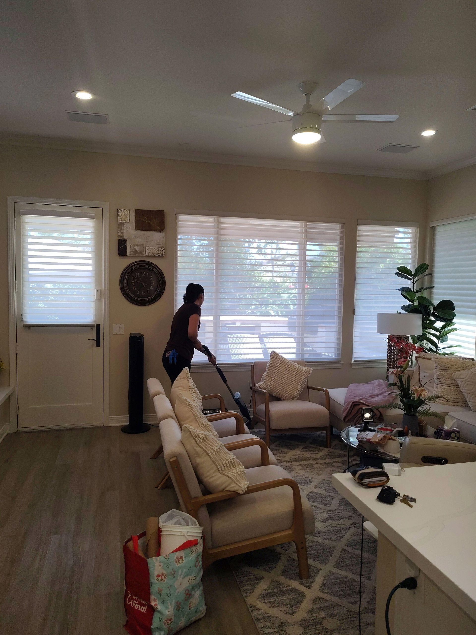 Living room with large windows, sofa and chairs; a person stands near the window beside a walker.