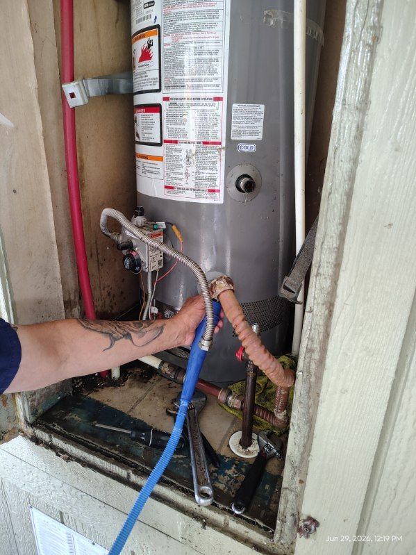 Water heater in a cramped closet with exposed, rusted and blue hoses being adjusted by a hand