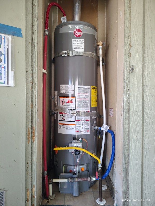 Gray water heater in a utility closet with exposed red, blue, and white pipes and warning labels.