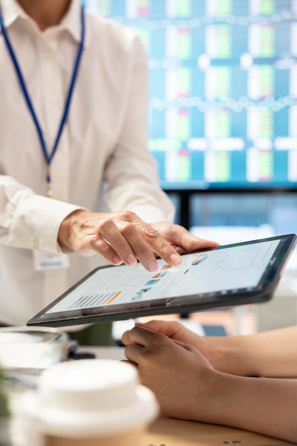 Person pointing at a tablet screen with graphs, another person's hands visible. In background, a monitor displays data.