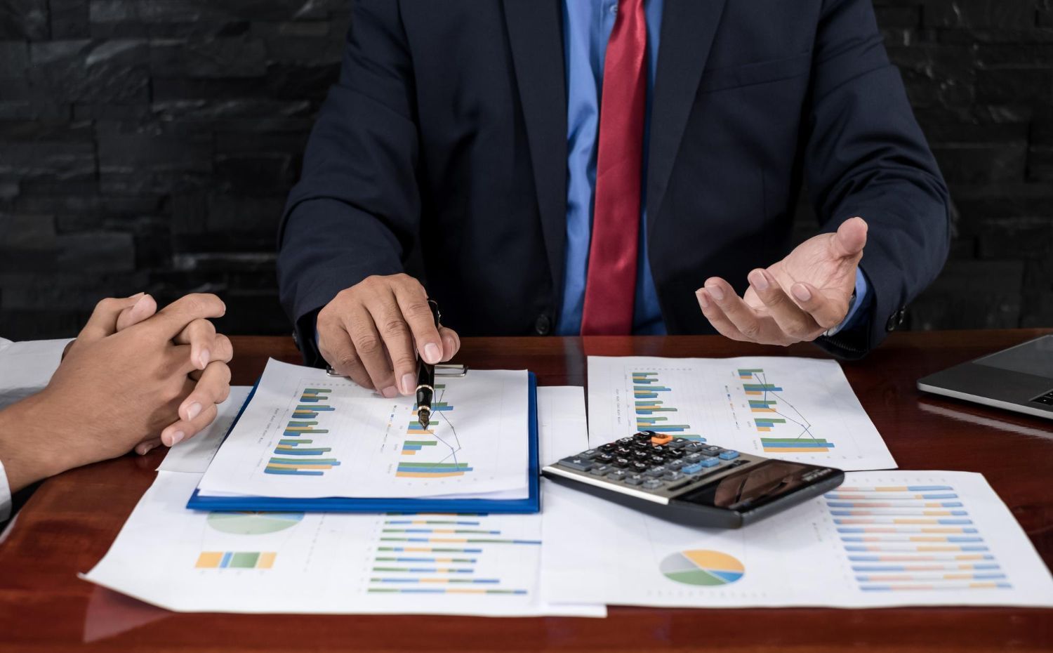 Man in suit pointing at charts during a meeting with a second person, a calculator is on the desk.