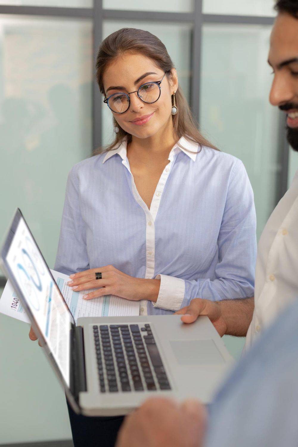 Woman in glasses looks at a laptop held by a person. Both are discussing a report, light setting.
