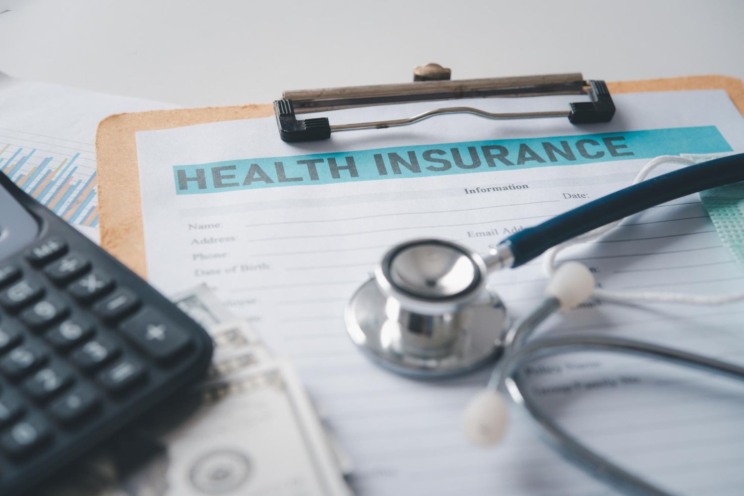 Health insurance form, stethoscope, calculator, and money on a wooden clipboard.