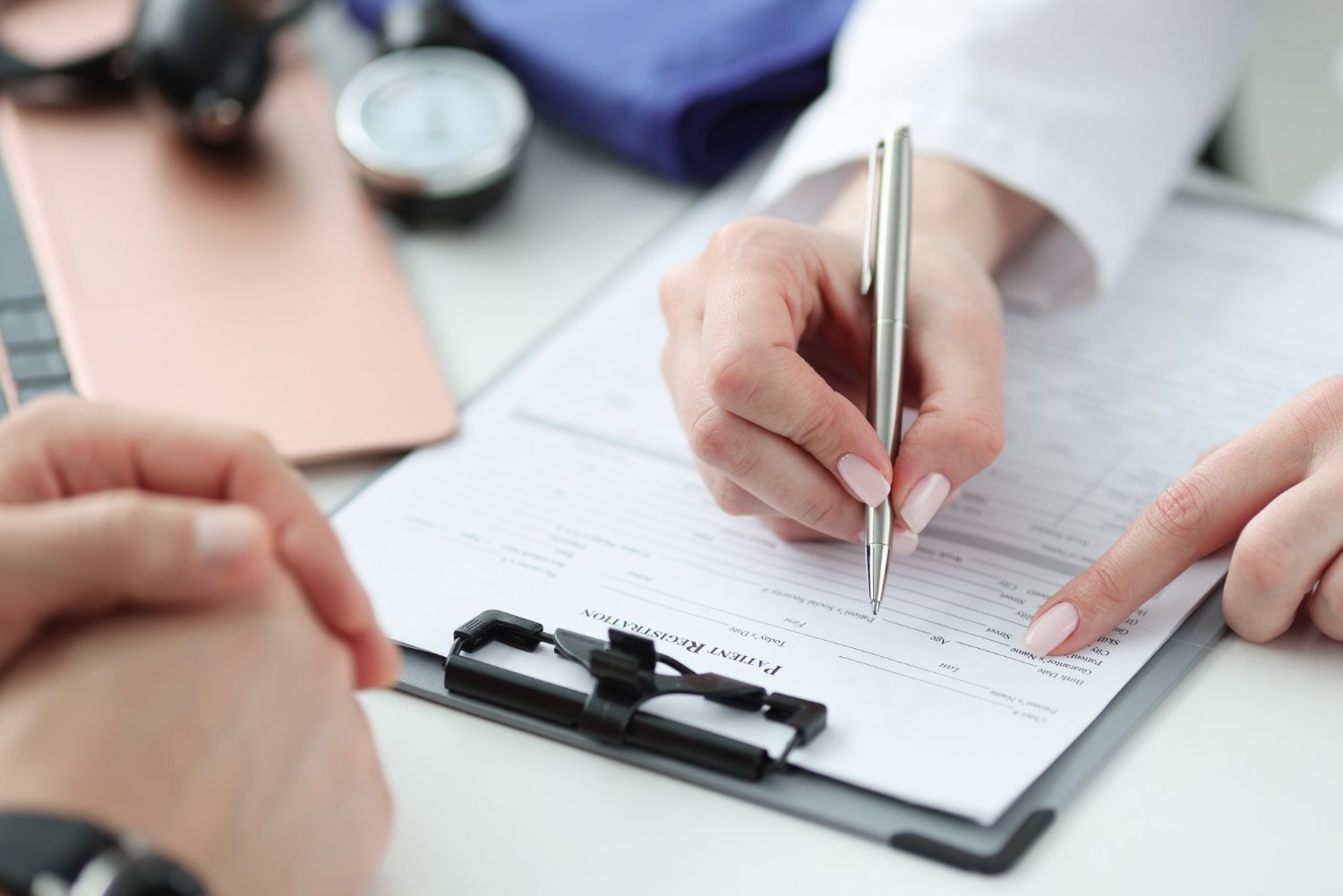 Doctor writing on a clipboard, pointing, with a patient in foreground and medical equipment in the background.