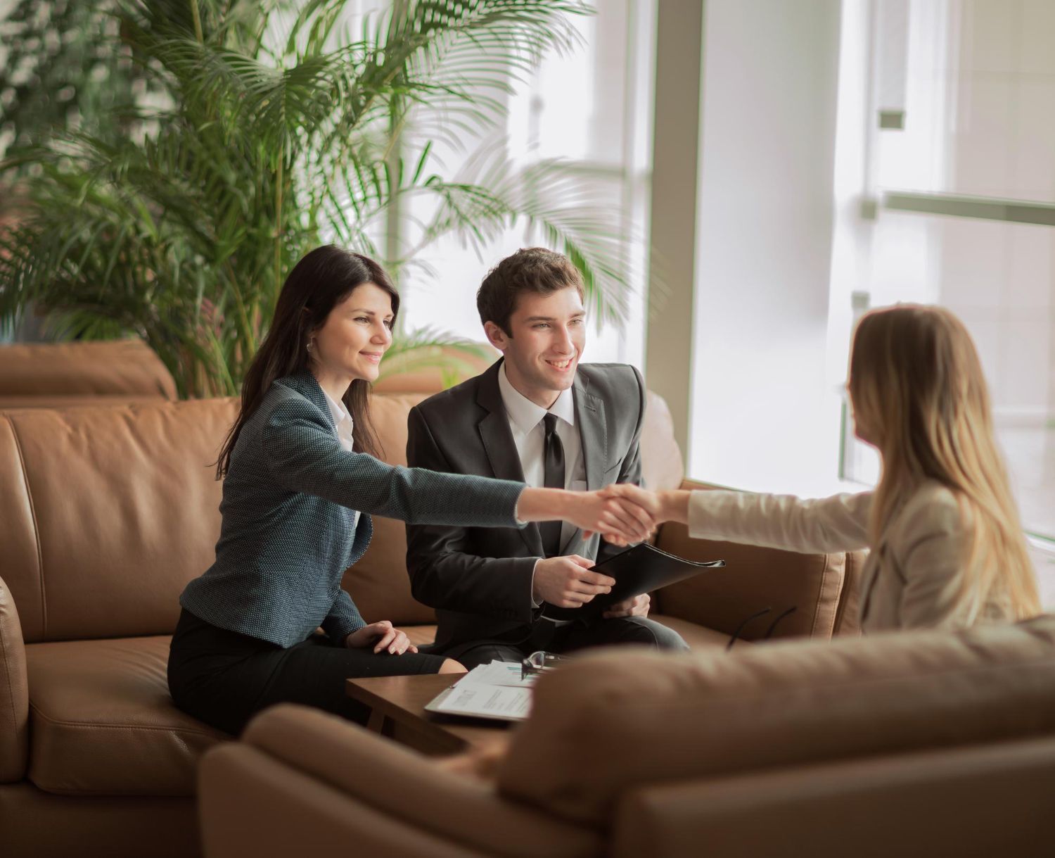 Business people shaking hands in a lobby, smiling. Leather sofa, greenery visible.