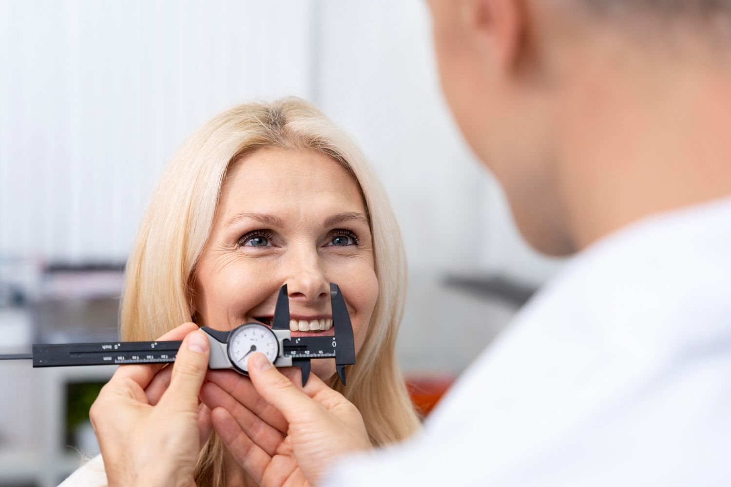 Person measuring a woman's nose with a caliper. The woman smiles. Indoors, medical setting.