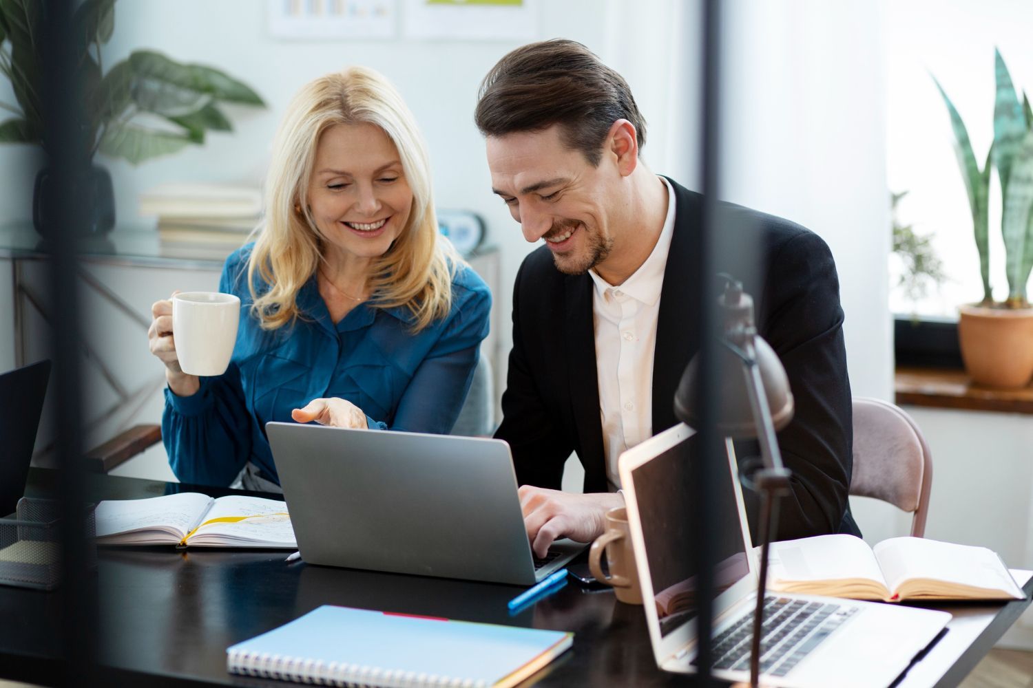 Two people at a desk looking at a laptop, smiling. Coffee cup, books, and plants visible.