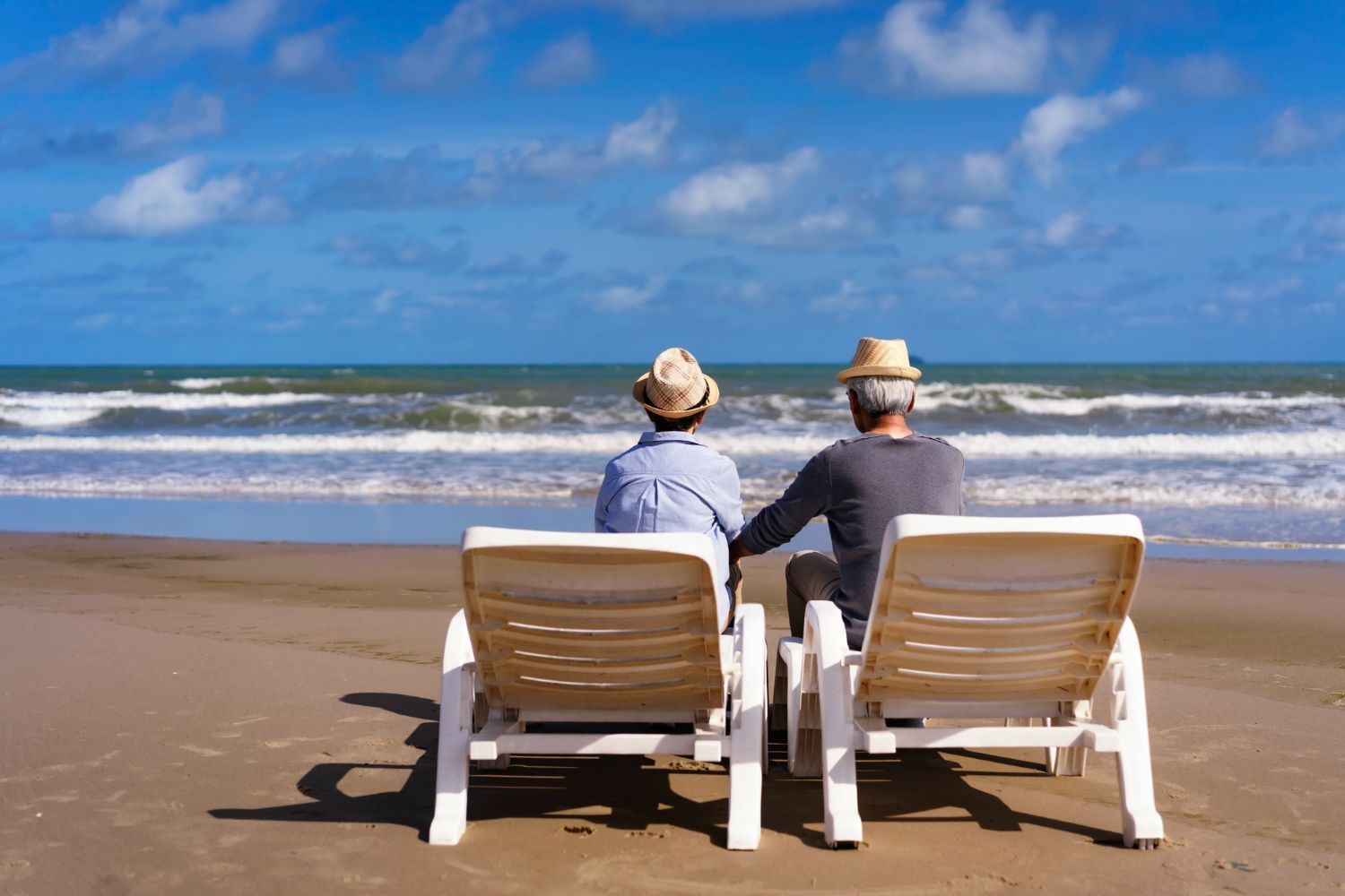 Two people relaxing in beach chairs, facing ocean under blue sky with clouds.