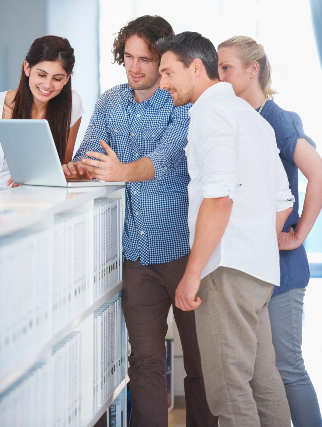 Four people gathered around a laptop, looking at the screen. They are in an office setting near a shelf of white binders.
