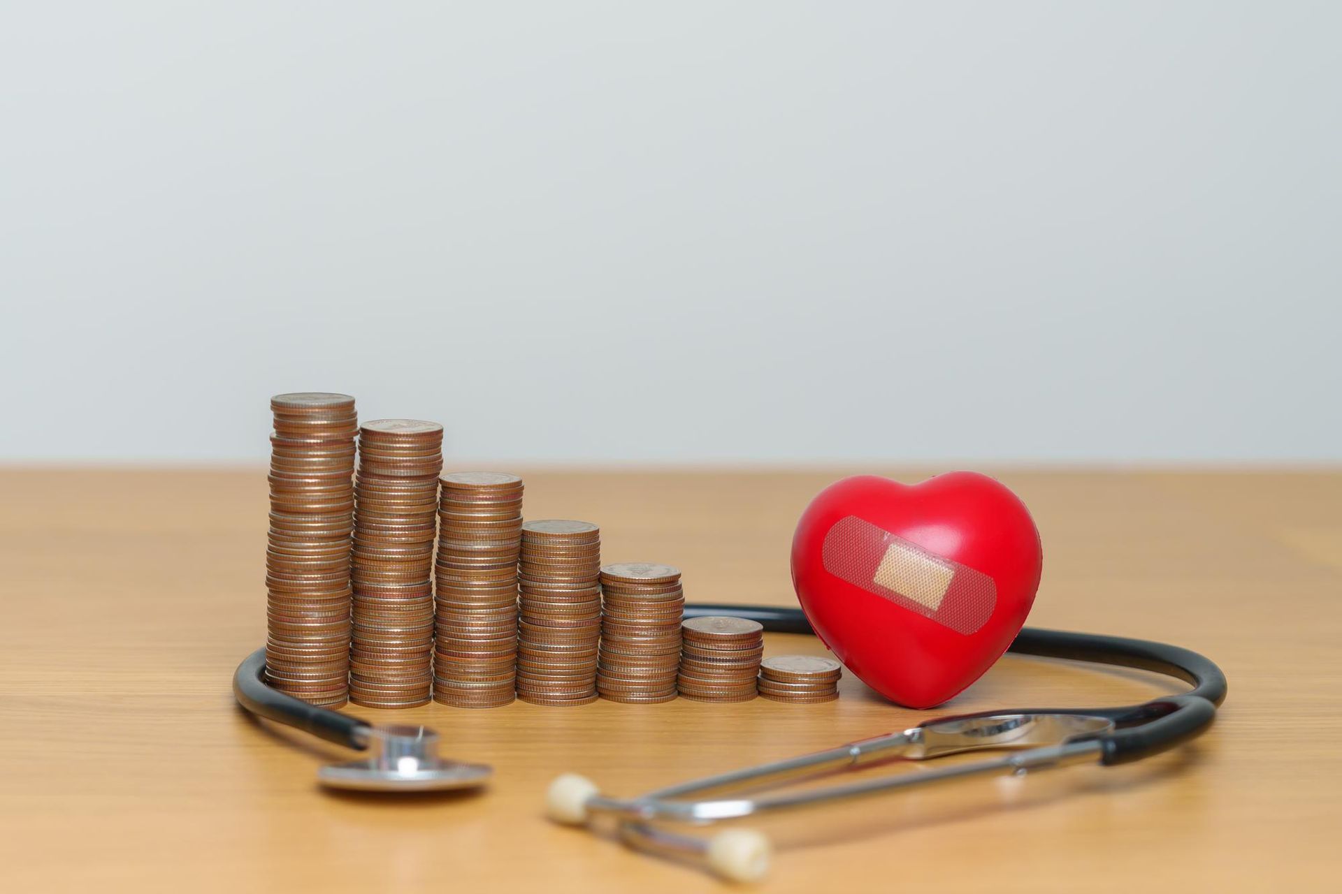 Stacks of coins, stethoscope, and red heart with bandage on wooden surface.