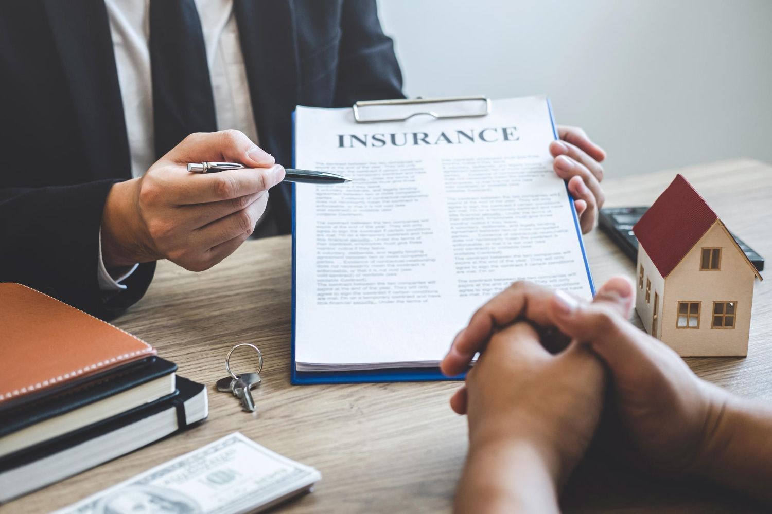 Person in suit pointing at insurance document, explaining to another person; miniature house, money, keys on table.