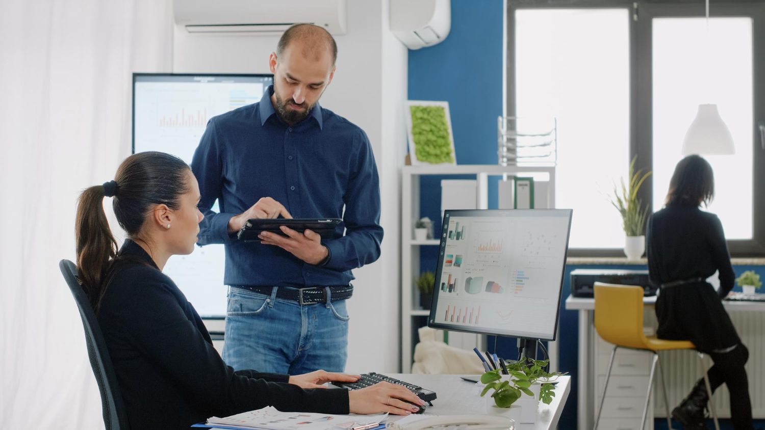 Woman seated at a desk, looking at a computer; man standing, holding a tablet; another person at a desk in the background.