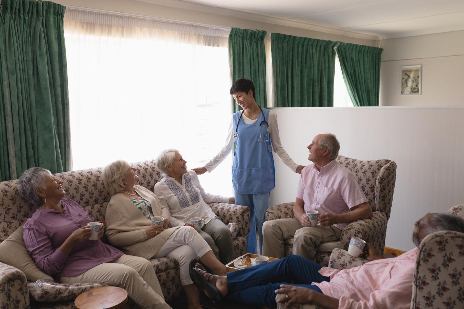 A caregiver in a blue uniform interacts with a group of seated residents in a living room setting.