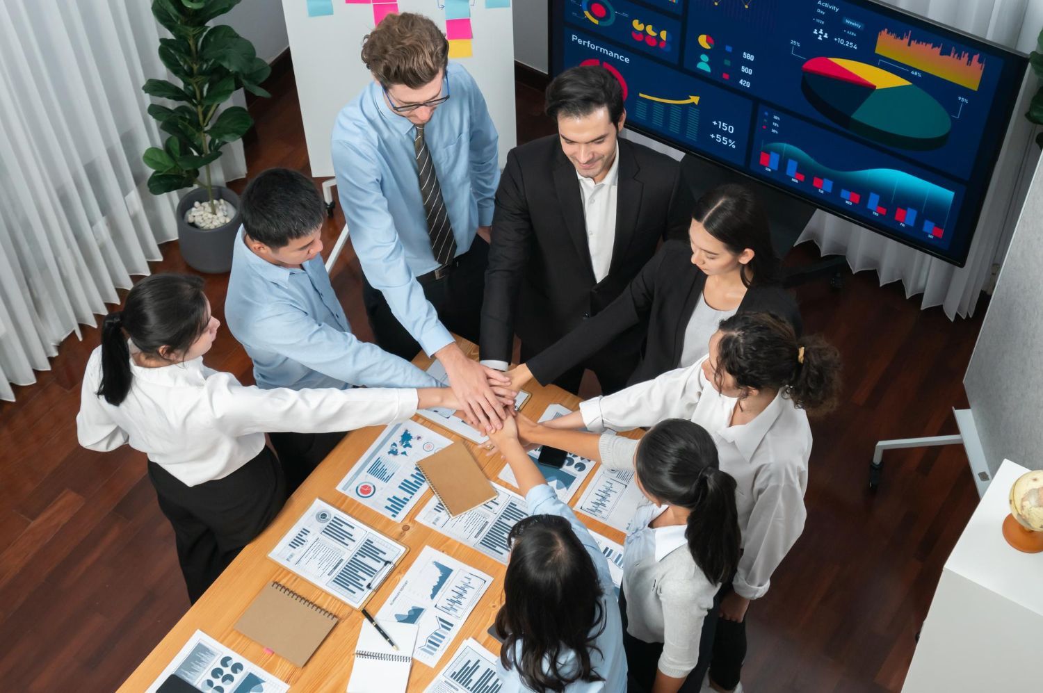 Team of professionals with hands stacked in a circle around a table covered in documents.