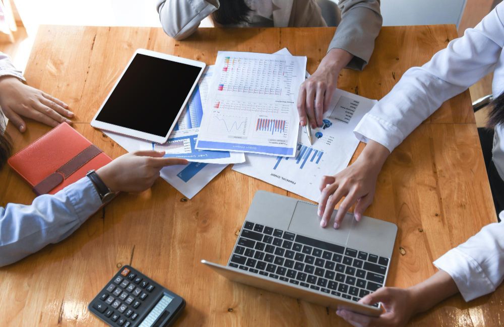 People collaborating at a wooden table reviewing documents, using a tablet, and laptop.