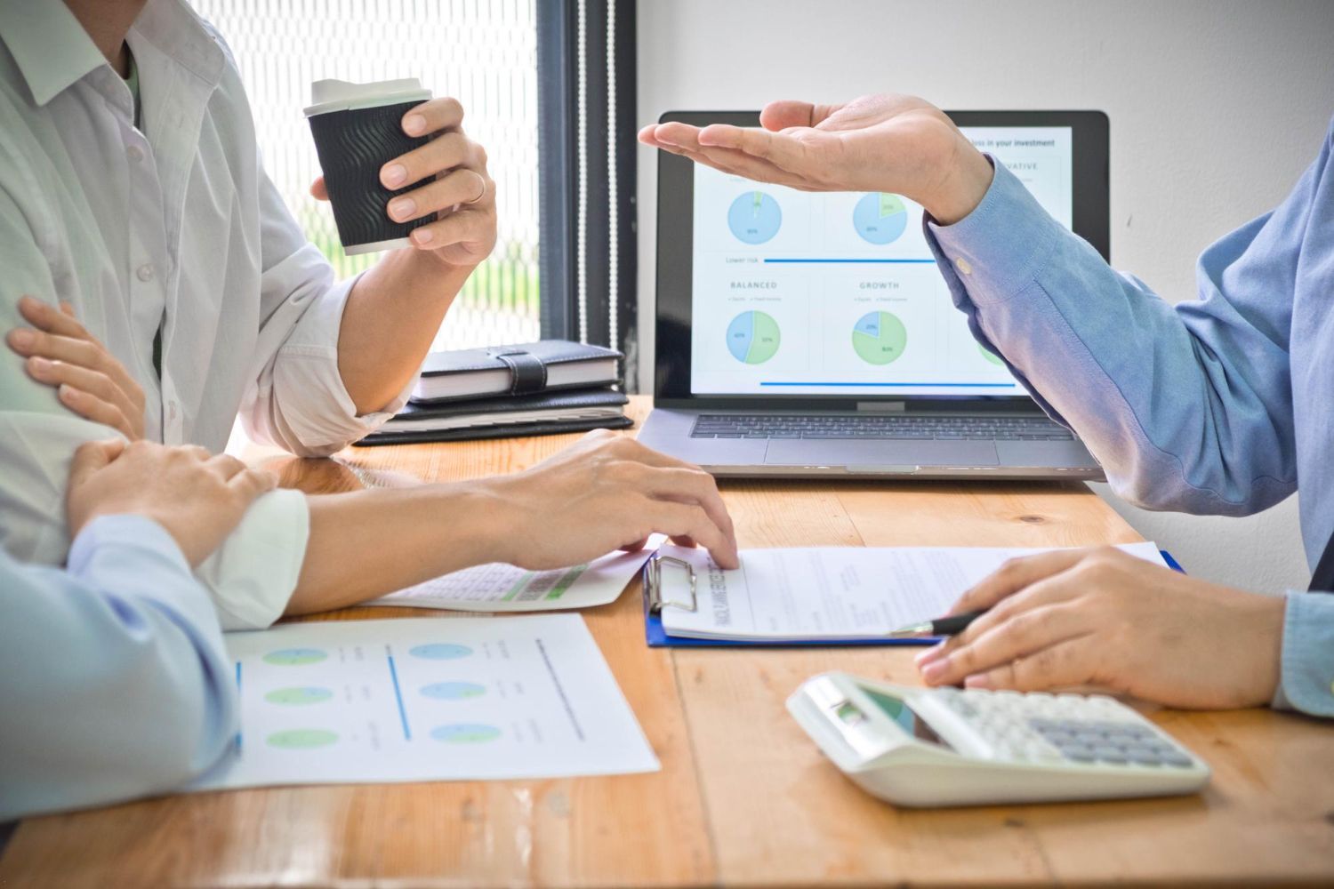People in business attire reviewing charts and documents at a table. One holds coffee, another gestures to a laptop screen.