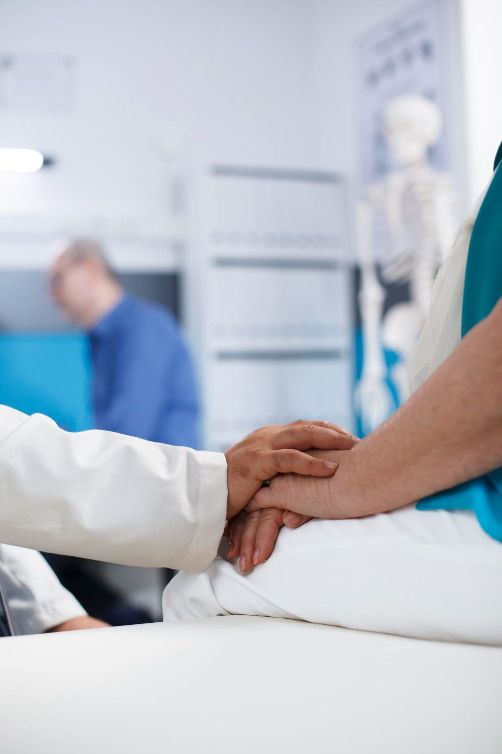 Doctor's hand on a patient's, offering comfort in a medical office; blurry background with skeleton model.