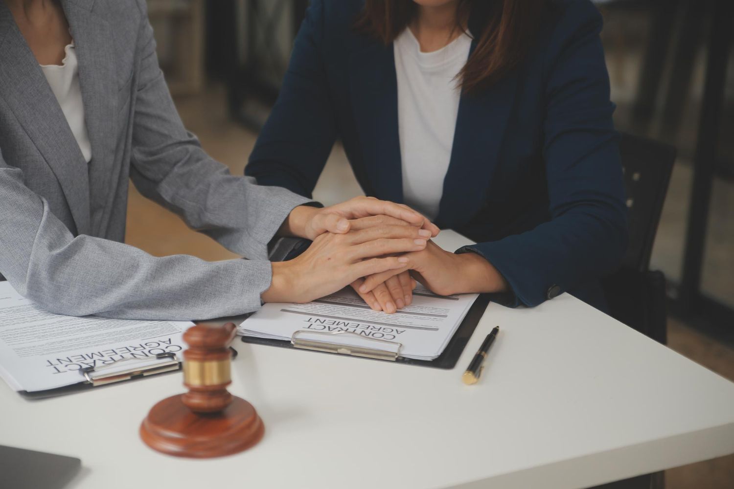 Two people's hands stacked on a table, with legal documents, a gavel, and a pen.