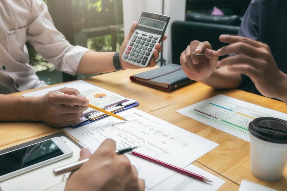People at a table review documents, calculator, smartphone, and coffee cup present, discussing finances.