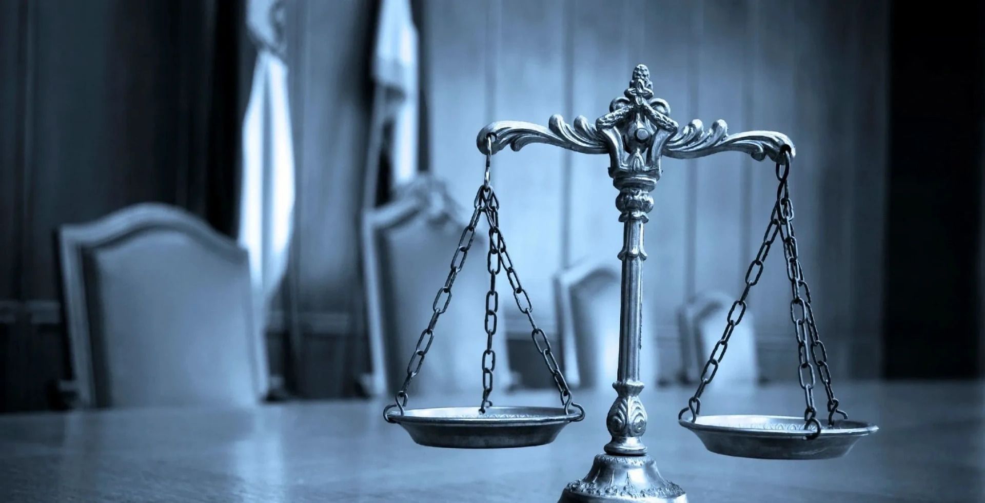 Scales of justice on a table in a courtroom with blurred chairs and a statue in the background.